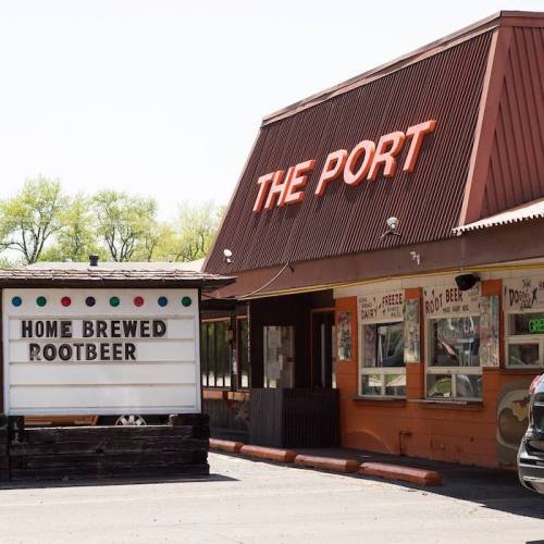 A roadside diner named "The Port" featuring a sign for home brewed root beer and colorful lights.