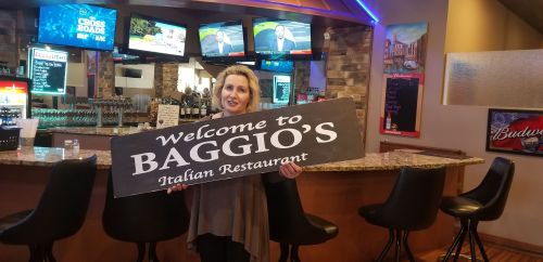 A woman holds a sign that reads "Welcome to Baggio's Italian Restaurant" in a bar area with TVs in the background.