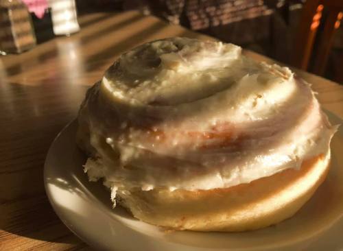 A close-up of a frosted cinnamon roll on a white plate, illuminated by warm sunlight.
