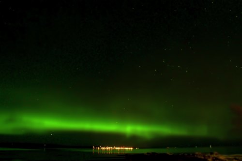 A vibrant green aurora borealis lights up the night sky over a calm body of water, with distant lights reflecting on the surface.