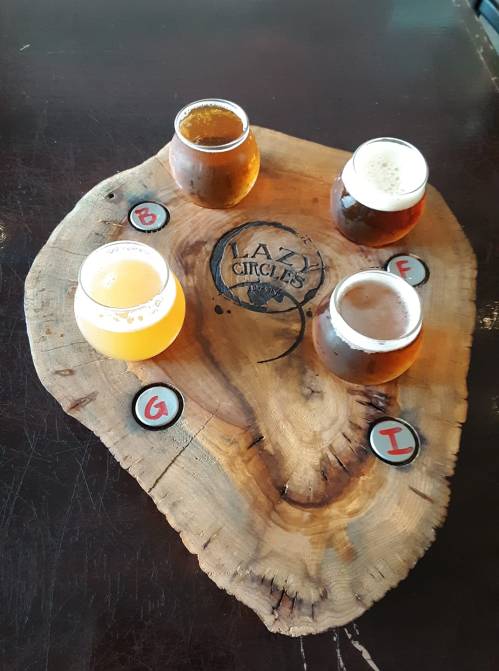 Four craft beer glasses on a wooden board with the logo "Lazy Circles" and labeled coasters.