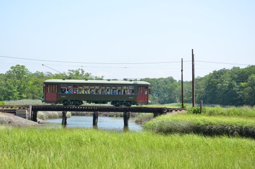A vintage train crosses a wooden bridge over a river, surrounded by lush green grass and trees under a clear blue sky.
