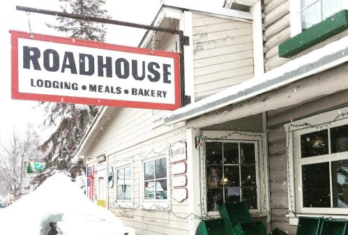 A snowy scene featuring a roadhouse sign, picnic table, and green benches outside a cozy lodge.