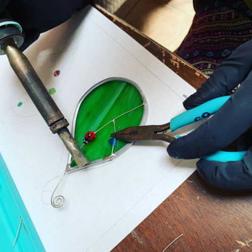 A person wearing gloves uses a soldering iron and pliers to work on a stained glass project featuring a green leaf and a ladybug.
