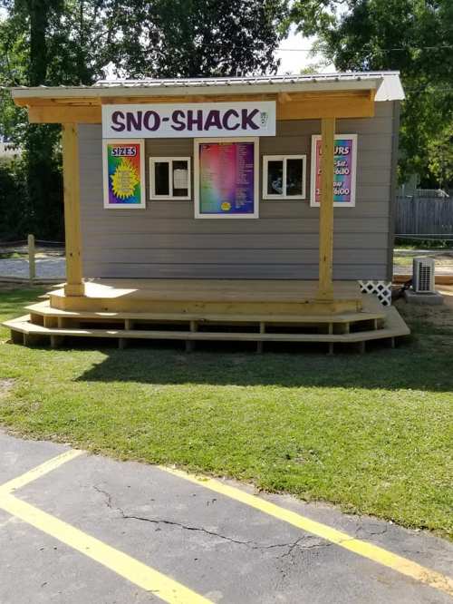 A small, wooden shack with a sign reading "Sno-Shack," featuring a menu and colorful decorations, set on a grassy area.
