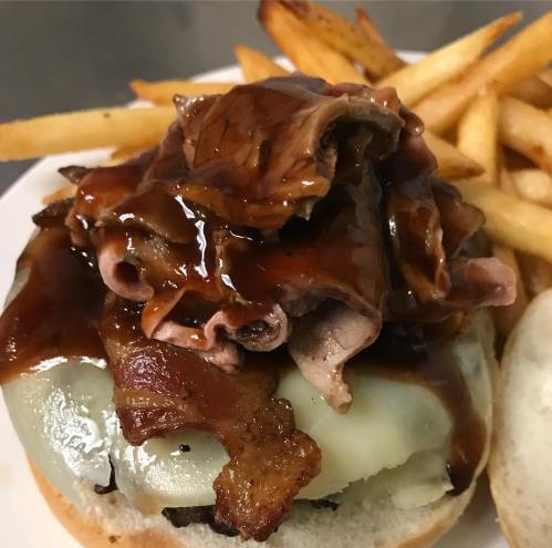 A close-up of a burger topped with melted cheese, sliced roast beef, bacon, and barbecue sauce, served with fries.