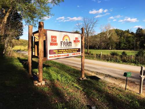 A roadside sign for "The Farm" with information about being open year-round, surrounded by trees and a grassy field.