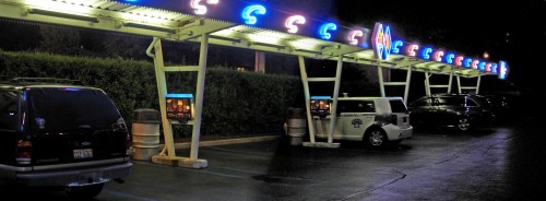 A nighttime view of a retro drive-in with neon lights and parked cars under illuminated canopies.