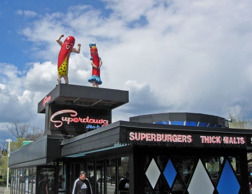 A drive-in restaurant with giant hot dog mascots on the roof, featuring a sign for "Superburgers" and "Thick Malts."