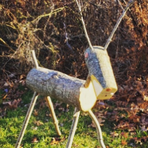 A wooden reindeer sculpture made from logs, set against a natural background with leaves on the ground.