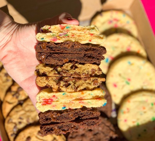 A hand holding a stack of colorful cookies, including chocolate chip and funfetti, with more cookies in the background.