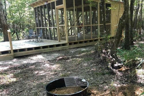 A wooden cabin with a screened porch sits in a wooded area, with two chairs on the deck and a fire pit nearby.