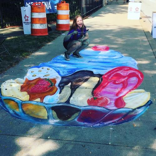 A girl poses next to a colorful 3D chalk drawing of a dessert plate on a sidewalk.