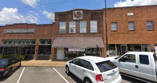 A brick building with a striped awning, featuring a "BREAKFAST" sign, flanked by shops and parked cars.
