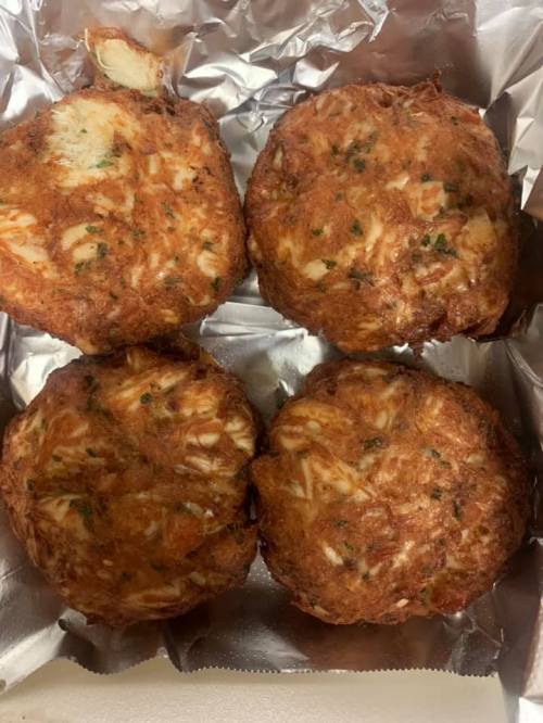 Four golden-brown crab cakes on a silver foil tray, garnished with green herbs.