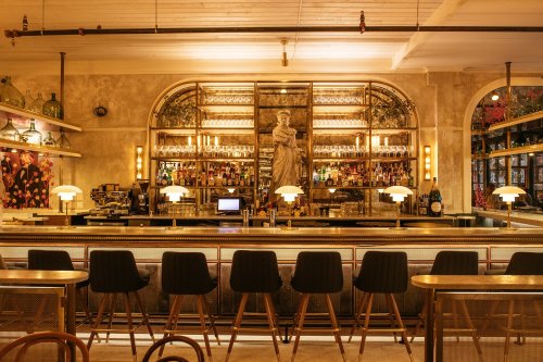 A stylish bar with a statue, shelves of bottles, and elegant lighting, featuring wooden stools and a warm ambiance.