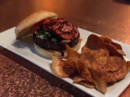 A juicy burger with lettuce, tomato, and bacon, served with a side of crispy potato chips on a white plate.