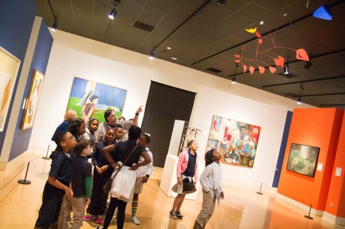 A group of children explores an art gallery, admiring colorful paintings and a mobile installation overhead.
