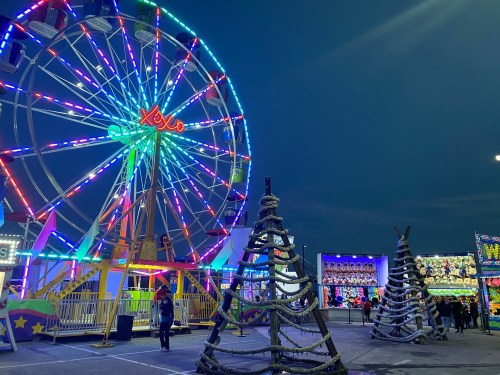 A brightly lit ferris wheel and festive Christmas trees at a nighttime fair, with colorful lights illuminating the scene.
