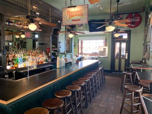 A cozy bar interior featuring a long counter, wooden stools, and shelves stocked with various drinks.