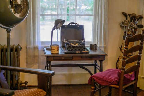 A vintage typewriter on a wooden desk beside a window, with a chair, lamp, and old-fashioned items in the room.