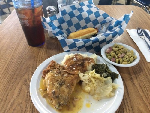 A plate of fried chicken with gravy, rice, collard greens, and cabbage, alongside a side of beans and a drink.