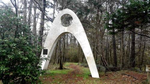 A large, white arch structure stands in a wooded area, surrounded by trees and greenery.