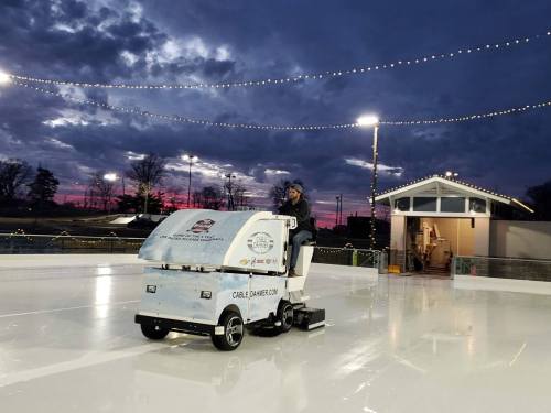 A person operates a Zamboni on an ice rink at dusk, with colorful lights and a sunset in the background.