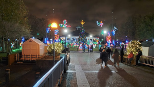 A festive park scene at night, featuring a large Christmas tree and colorful holiday lights, with people walking around.