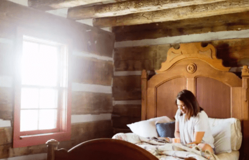 A woman sits on a bed in a rustic room with wooden walls, reading in soft, natural light from a nearby window.