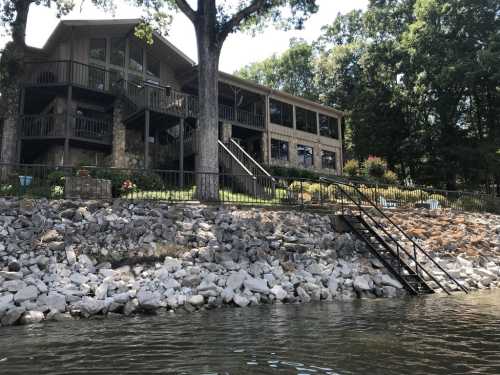 A lakeside view of a large wooden house with a stone foundation, surrounded by trees and a rocky shoreline.