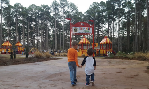Two children walk hand in hand towards a colorful playground surrounded by tall trees.