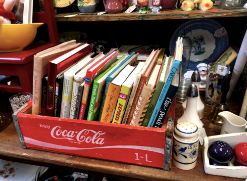 A vintage Coca-Cola crate holds a collection of colorful cookbooks on a wooden shelf surrounded by various knick-knacks.