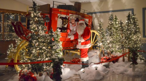 Santa Claus and a helper pose by a Wells Fargo stagecoach, surrounded by festive trees and holiday decorations.