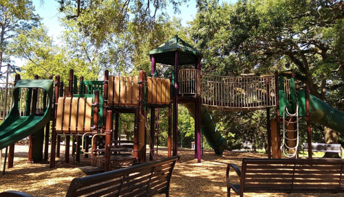 Colorful playground structure with slides and climbing areas, surrounded by trees and benches in a sunny park.