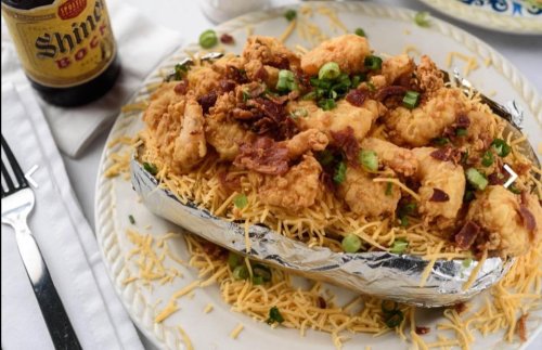 A loaded dish with crispy chicken, bacon, and green onions on a bed of shredded cheese, served in a foil tray.