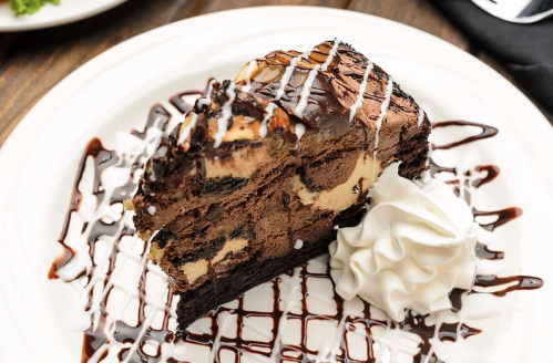 A slice of rich chocolate cake drizzled with chocolate sauce and topped with whipped cream on a white plate.