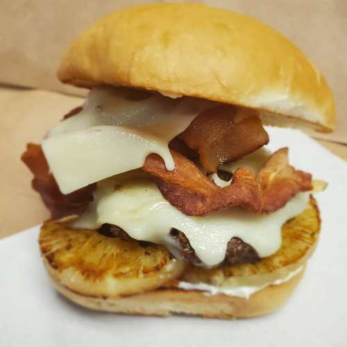 A stacked burger with bacon, melted cheese, grilled pineapple, and a soft bun on a white napkin.