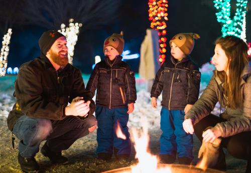 A family gathers around a fire, enjoying a winter evening with colorful lights in the background. Two children wear hats.
