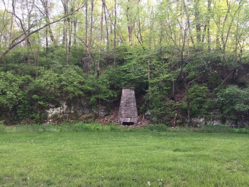 A stone chimney stands against a lush green hillside, surrounded by trees and grass.
