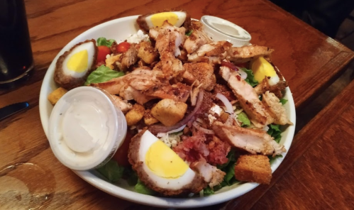 A hearty salad topped with grilled chicken, croutons, sliced eggs, and dressing on a wooden table.