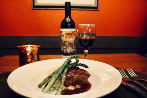 A plate of steak with asparagus, mashed potatoes, a glass of red wine, and a candle on a table with a red wall backdrop.