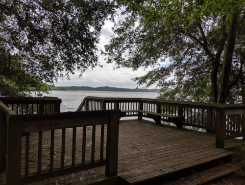 A wooden deck overlooking a calm lake, surrounded by trees under a cloudy sky.