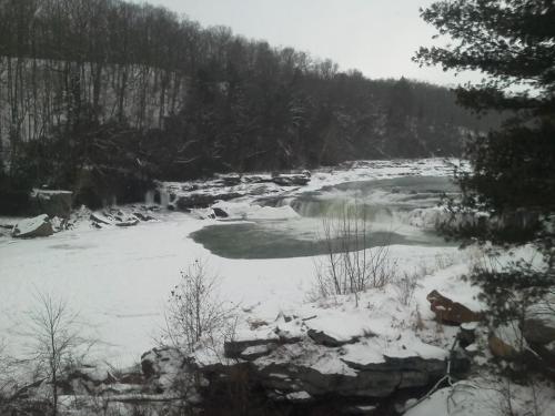 A snowy landscape featuring a river with flowing water and rocky banks, surrounded by trees and a wintery backdrop.