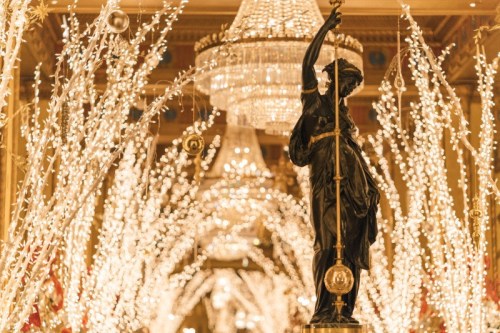 A beautifully lit hallway adorned with twinkling lights and a statue holding a staff, under elegant chandeliers.