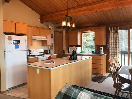 Cozy kitchen with wooden cabinets, island, and dining area, featuring large windows and warm lighting.