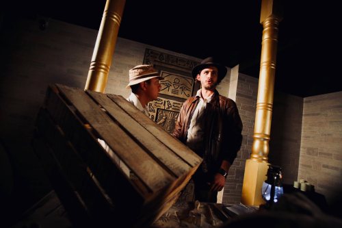 Two men in a dimly lit room with ancient Egyptian decor, one holding a wooden crate and the other wearing a hat.