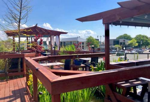A sunny outdoor deck with wooden structures, greenery, and a view of a nearby street and buildings.