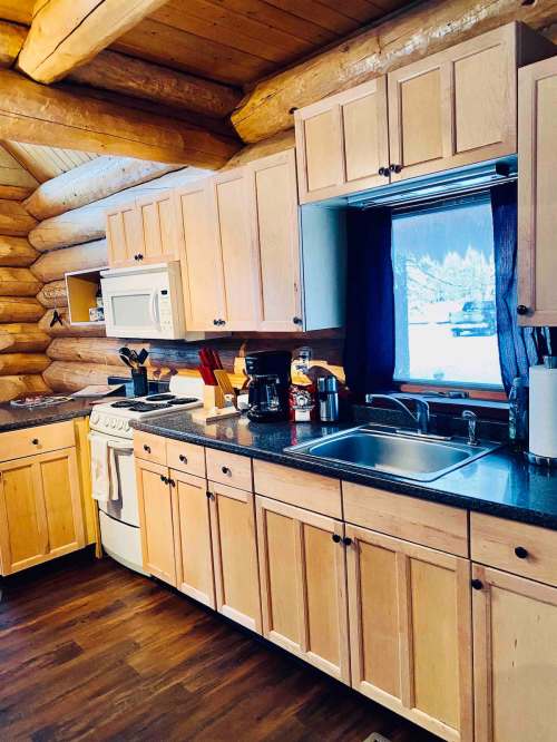 A cozy log cabin kitchen with wooden cabinets, a sink, stove, and a window overlooking a snowy landscape.