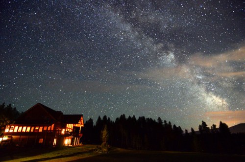 A cozy cabin illuminated at night under a starry sky filled with the Milky Way and silhouetted trees.